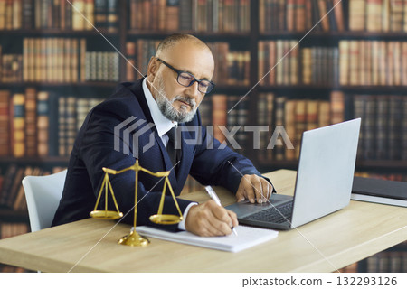 Senior lawyer working at laptop with scales of justice in law library office Senior lawyer working at laptop with scales of justice in law library office 132293126