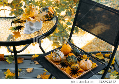 White decorative skull with maple leaves and mixed gourds on a glass table and chair on autumn balcony. Modern spooky decor, visual Halloween culture, alternative seasonal styling, aesthetic horror White decorative skull with maple leaves and mixed gourds on a glass table and chair on autumn balcony. Modern spooky decor, visual Halloween culture, alternative seasonal styling, aesthetic horror 132293812