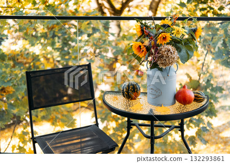 Simple autumn balcony setup with floral centerpiece in a vintage can beside gourds and a mesh chair against fall foliage. Compact seasonal design, minimalist outdoor decor, cozy urban living 132293861