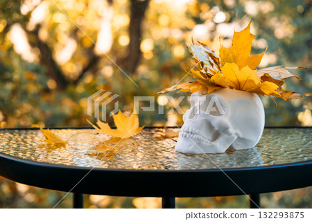 White skull planter topped with yellow maple leaves on a glass table against a blurred autumn background. Halloween aesthetics, seasonal decor minimalism, spooky chic, modern horror styling 132293875