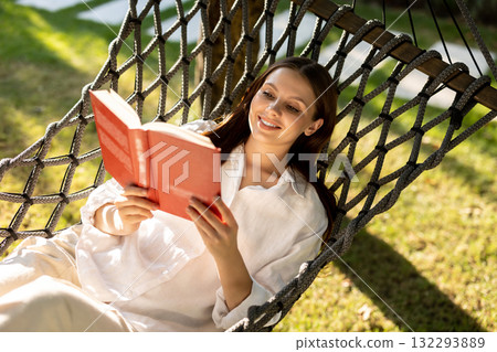 Long-haired caucasian young woman enjoying leisure while lying in hammock with a book 132293889