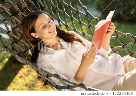 Young woman lying in hammock, reading a book and looking contented 132294368