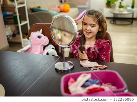 Little girl enjoys grooming her hair while seated at a cozy table adorned with colorful accessories and adorable toys 132296090