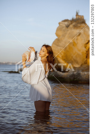 Young woman in summer white dress holds big shell in her hands Young woman in summer white dress holds big shell in her hands 132297803