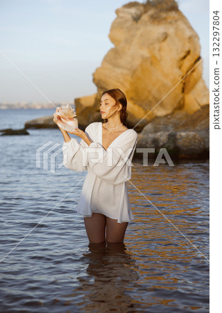Young woman in summer white dress holds big shell in her hands 132297804