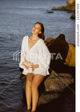 Young woman in summer white dress sits on a rock in the sea in her hands she holds a shell Young woman in summer white dress sits on a rock in the sea in her hands she holds a shell 132297809