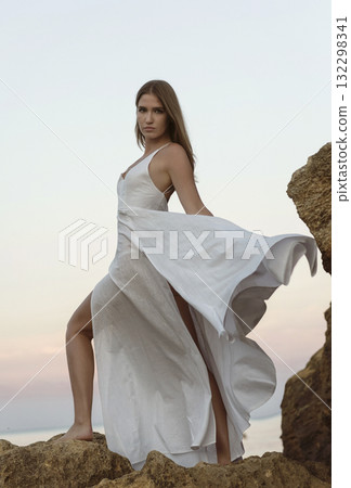 young girl in a long white dress posing on a cliff against the backdrop of the sea and sunset sky young girl in a long white dress posing on a cliff against the backdrop of the sea and sunset sky 132298341