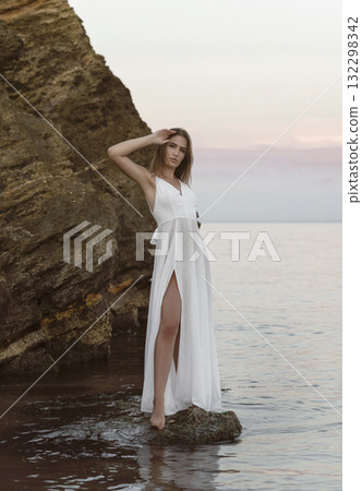 young girl in a long white dress poses on a rock against the backdrop of a calm sea and sunset sky young girl in a long white dress poses on a rock against the backdrop of a calm sea and sunset sky 132298342