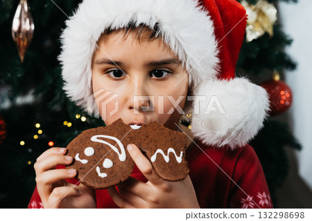 Christmas Portrait of Kid with Gingerbread and Holiday Decor. Child wearing a Santa hat and red pajamas eating gingerbread cookies in front of a decorated Christmas tree with lights and ornaments Christmas Portrait of Kid with Gingerbread and Holiday Decor. Child wearing a Santa hat and red pajamas eating gingerbread cookies in front of a decorated Christmas tree with lights and ornaments 132298698