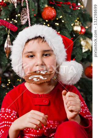 Christmas Portrait of Kid with Gingerbread and Holiday Decor. Child wearing a Santa hat and red pajamas eating gingerbread cookies in front of a decorated Christmas tree with lights and ornaments Christmas Portrait of Kid with Gingerbread and Holiday Decor. Child wearing a Santa hat and red pajamas eating gingerbread cookies in front of a decorated Christmas tree with lights and ornaments 132298699