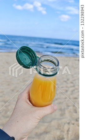 Hand holding a bottle of orange juice against a sunny beach and blue ocean background 132298943
