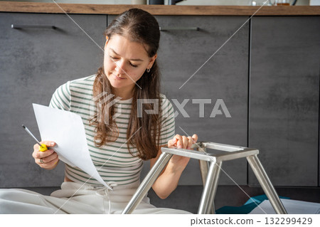 Smiling young woman sitting on the floor reading instructions while assembling a chair. Continuation of the series showing joy and satisfaction from independent DIY work at home. 132299429