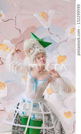 Smiling rococo woman with lollipop and green paper hat posing on floral backdrop 132299599