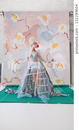 Playful woman blowing pink bubblegum in front of floral backdrop in character of Marie Antoinette. 132299604