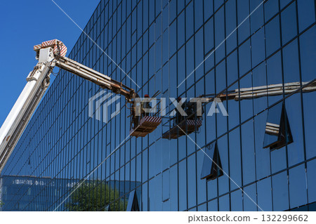 A construction crane with a cradle or lifting platform is reflected in the facade glazing of an office building. A construction worker in the crane bucket works on the facade glazing. 132299662