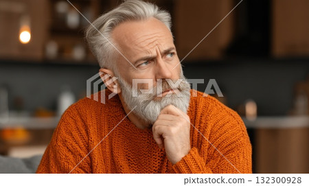 Mature man with grey hair wearing an orange sweater looks pensive while sitting at home Mature man with grey hair wearing an orange sweater looks pensive while sitting at home 132300928