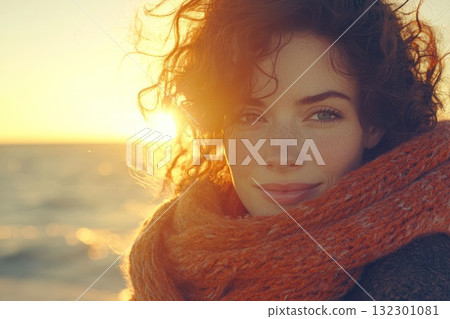 A woman with curly hair is smiling while wearing a cozy scarf during sunset by the beach 132301081
