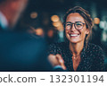 A woman with glasses smiles warmly while shaking hands with a man in a modern workspace 132301194