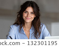 Young woman seated at desk with a laptop smiling while holding a pen and engaging in work tasks 132301195