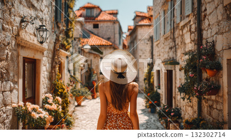Woman in summer dress and wide brimmed hat walking through charming old stone street with flowers, feeling relaxed and joyful 132302714
