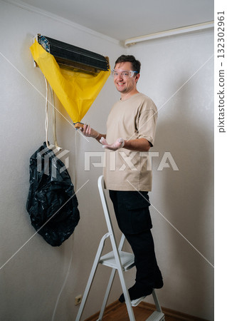 Vertical portrait of positive technician cleaning air conditioning unit, wearing protective gear, using sponge, yellow cover, smiling looking at camera. Concept of air condition hygiene. Vertical portrait of positive technician cleaning air conditioning unit, wearing protective gear, using sponge, yellow cover, smiling looking at camera. Concept of air condition hygiene. 132302961