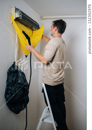 Vertical shot of technician male cleaning air conditioning unit using steam cleaner with protective sheet catching debris. Concept for maintenance of hygiene and pure air to prevent allergies 132302976