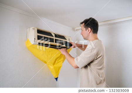 Technician standing on ladder, cleaning air conditioner unit using portable vacuum cleaner and protective bag to prevent water from dripping on floor during maintenance. Technician standing on ladder, cleaning air conditioner unit using portable vacuum cleaner and protective bag to prevent water from dripping on floor during maintenance. 132303009