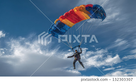 Skydiver with colorful parachute soaring through dramatic cloudy sky, extreme sport adventure with vibrant red, orange and blue canopy against clouds 132303307