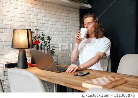 Man sipping coffee while working on laptop at a cozy desk during daytime Man sipping coffee while working on laptop at a cozy desk during daytime 132303876