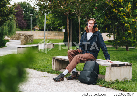 Young man relaxing in park while listening to music on a sunny day with a backpack nearby 132303897