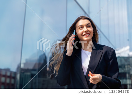 Young Woman Smiling While Talking on Her Phone in an Urban Setting Near a Glass Building on a Sunny Day Young Woman Smiling While Talking on Her Phone in an Urban Setting Near a Glass Building on a Sunny Day 132304378