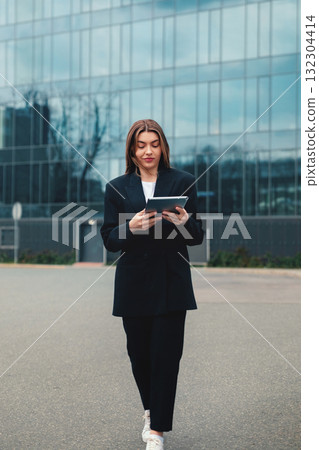 Young Adult Walks Confidently While Using a Tablet Outside a Modern Office Building in an Urban Area During the Daytime Young Adult Walks Confidently While Using a Tablet Outside a Modern Office Building in an Urban Area During the Daytime 132304414