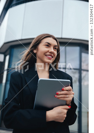 Woman Holding a Tablet While Standing Outside a Modern Office Building on a Sunny Day With a Cheerful Expression Woman Holding a Tablet While Standing Outside a Modern Office Building on a Sunny Day With a Cheerful Expression 132304417