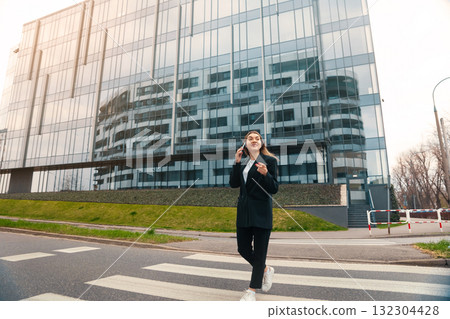 Young Woman Walks Confidently Across a Pedestrian Crosswalk in an Urban Setting During Daylight, Reflecting Modern Architecture of Nearby Buildings 132304428