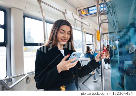 Young Woman Enjoys Texting on Her Smartphone While Riding a City Tram in the Afternoon, Surrounded by Fellow Commuters and Urban Scenery Young Woman Enjoys Texting on Her Smartphone While Riding a City Tram in the Afternoon, Surrounded by Fellow Commuters and Urban Scenery 132304434