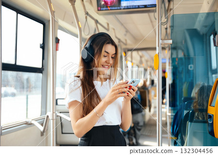 Young Woman Smiles While Using Smartphone on Public Transport With Headphones During Daytime Commute 132304456