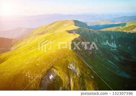Aerial view of rugged mountain range with lush green slopes at sunset. Landscape showcases beauty of untouched nature with distant peaks and valleys bathed in soft sunlight. Carpathians, Chornogora. 132307396