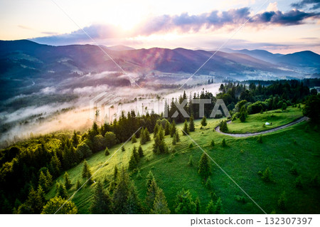 Breathtaking aerial view of lush green valley at sunrise, where morning mist gracefully floats above landscape. Sun's rays cast golden glow, creating tranquil and picturesque scene. 132307397