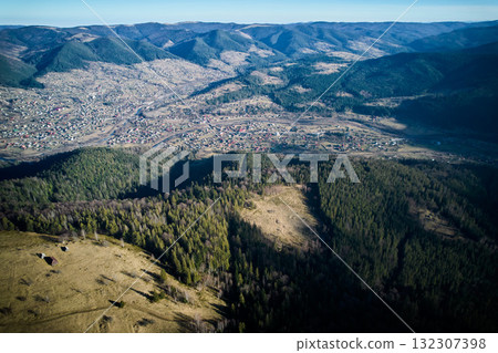Aerial view of mountain landscape with sprawling village nestled in valley. Rolling hills dense forests create picturesque scene under clear blue sky. Natural beauty and tranquility of the area. 132307398