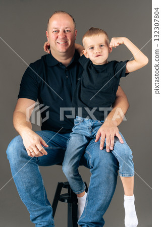 Father holding son on lap, boy showing muscles joyful family studio portrait expressing pride and happiness Father holding son on lap, boy showing muscles joyful family studio portrait expressing pride and happiness 132307804