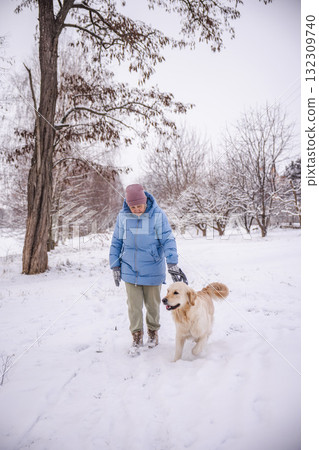 Elderly woman in a blue winter coat walking her Golden Retriever through a snowy rural landscape. The dog walks happily beside her as she enjoys a quiet moment outdoors surrounded by winter trees. 132309740