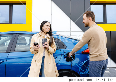 Young couple man and woman traveling by electric car having stop at charging station. Boyfriend plugging in cable to charge. Man talking with girlfriend, holding cup drinking hot coffee smiling Young couple man and woman traveling by electric car having stop at charging station. Boyfriend plugging in cable to charge. Man talking with girlfriend, holding cup drinking hot coffee smiling 132310158