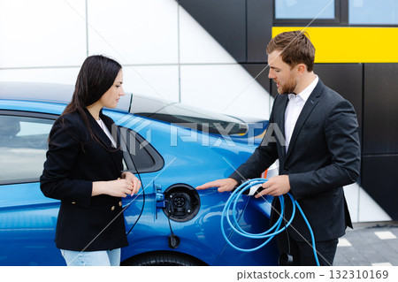 Sales manager showing how to charge electric car to a young client woman, plugging wire into the car socket at the car dealership outdoor 132310169