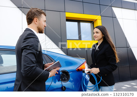 Sales manager showing how to charge electric car to a young client woman, plugging wire into the car socket at the car dealership outdoor Sales manager showing how to charge electric car to a young client woman, plugging wire into the car socket at the car dealership outdoor 132310170