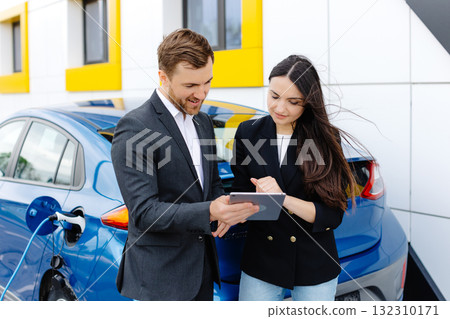 Smiling woman standing at the car and pointing at car seller tablet. She is picked right car for her she saw online. A woman concludes an agreement to buy a car and shakes the manager's hand 132310171