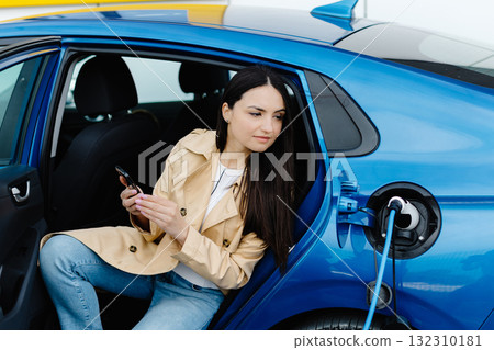 Business lady sitting inside charging electric car and using modern smartphone. Technology and transport concept. Business lady sitting inside charging electric car and using modern smartphone. Technology and transport concept. 132310181