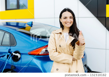Woman is standing at the car and charging electric car at the charging station and and showing finger up gesture Woman is standing at the car and charging electric car at the charging station and and showing finger up gesture 132310182