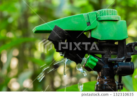 A green sprinkler head is positioned among lush green plants. Water sprays from the nozzle, demonstrating an irrigation system in action. A green sprinkler head is positioned among lush green plants. Water sprays from the nozzle, demonstrating an irrigation system in action. 132310635