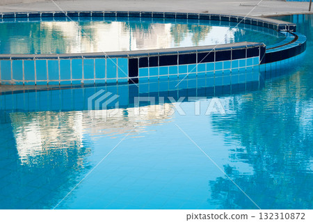 A serene swimming pool with clear blue water, surrounded by palm trees and landscaped gardens. The pool features a curved design and tiled edges. 132310872