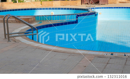 An empty swimming pool with blue tiles and a ladder. The water is absent, revealing the tiled bottom. Surrounding greenery is visible in the background. 132311016